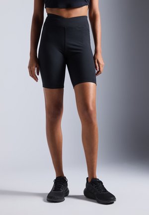 Model draagt zwarte hoge taille fietsbroek, zwarte cropped top en zwarte sportschoenen, staand tegen een grijze verloopachtergrond.