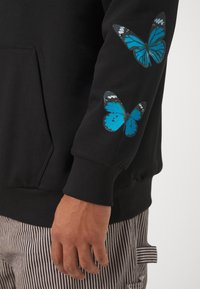 Černá mikina s kapucí s dvěma modrými potisky motýlů na pravém rukávu, vyrobená z měkké látky s volným střihem a žebrovanými manžetami.