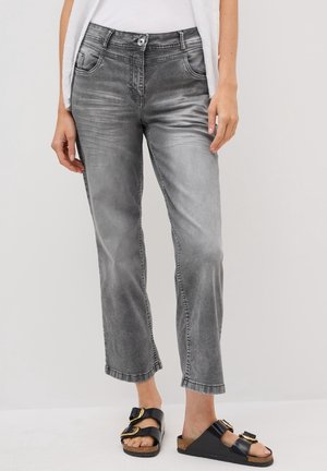 Femme portant un jean droit gris délavé et des sandales noires slip-on avec des boucles dorées, debout devant un fond clair uni.