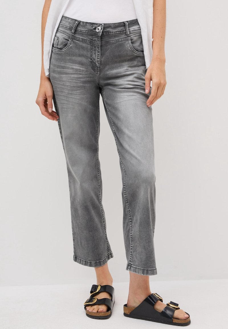 Femme portant un jean droit gris délavé et des sandales noires slip-on avec des boucles dorées, debout devant un fond clair uni.