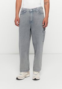 Grijze denim cargo broek met een losse pasvorm, voorzien van een grote zijzak, vijf riemlussen en een knoopsluiting. Draagt met beige sneakers.