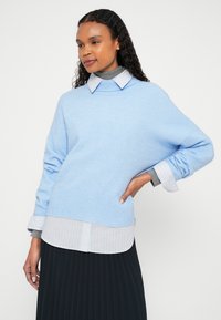 Svetlomodrý pletený sveter s okrúhlym výstrihom, spustenými ramenami a rebrovaným lemom, vrstvený cez bielu košeľu s jemnými pruhmi a tmavú plisovanú sukňu.