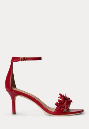 Κόκκινο δερμάτινο σανδάλι με ψηλό τακούνι, λουράκι αστραγάλου και λουλουδάτη διακόσμηση στο λουράκι των δαχτύλων, εμφανίζεται σε λευκό φόντο.