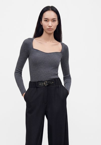 Femme aux longs cheveux noirs portant un pull gris ajusté à col carré et un pantalon noir taille haute avec ceinture, mains dans les poches, fond neutre.