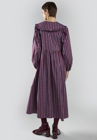 Vestido largo de cuadros burdeos con cuello volante y mangas abullonadas; cuenta con una cintura ajustada y una falda acampanada. Combinado con zapatos burdeos brillantes.