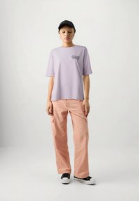 Ung person iført en sort cap, løs lyserød t-shirt med lille grafik, ferskenfarvede cargo-bukser og sort-hvide sneakers, stående mod en hvid baggrund.