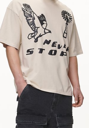 Camiseta beige con gráfica negra de un pájaro volando, una flor y el texto "NUNCA PARES", combinada con pantalones cargo negros.