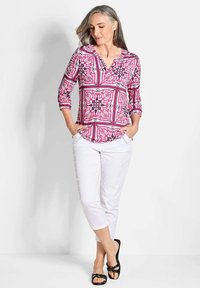 Rosa gemusterte Bluse mit ¾-Ärmeln, V-Ausschnitt, floralen Design, kombiniert mit weißen Caprihosen und schwarzen Sandalen.