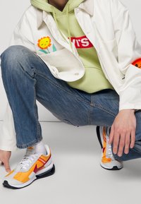 Veste blanche à motif floral, sweat à capuche vert, jean bleu et baskets multicolores avec des accents blancs, orange, jaunes et rouges.