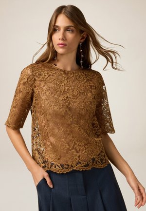 Top in pizzo di colore marrone ricco con motivi floreali, maniche corte e orlo a scallop. Indossato sopra una gonna plissettata scura, che mette in mostra una texture trasparente.
