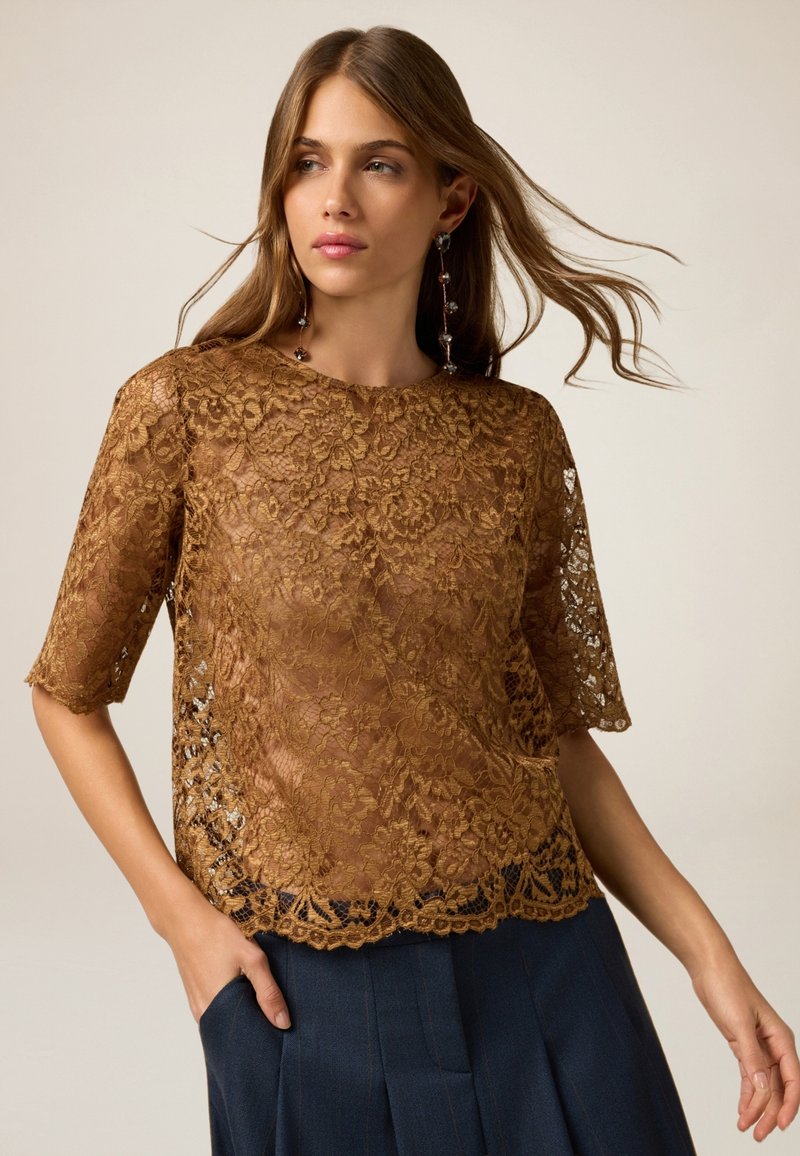 Top in pizzo di colore marrone ricco con motivi floreali, maniche corte e orlo a scallop. Indossato sopra una gonna plissettata scura, che mette in mostra una texture trasparente.