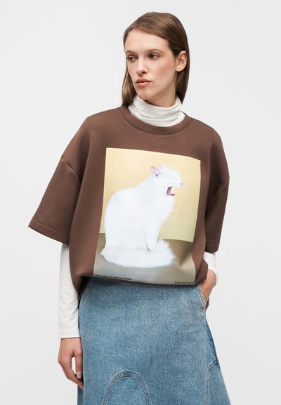 Bruine oversized sweatshirt met een witte kattenafbeelding op een gouden achtergrond. Model draagt het over een witte coltrui, gecombineerd met een denim rok.