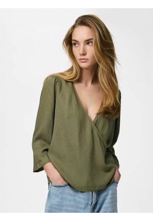 Kobieta o długich blond włosach, ubrana w oliwkowo-zieloną bluzkę kopertową i jasnoniebieskie jeansy, stoi z rękami w kieszeniach, patrząc na bok.