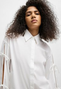White button-up shirt with long sleeves featuring open side details tied with bows. Subtle “HUGO” logo embroidered on the chest. Smooth texture.