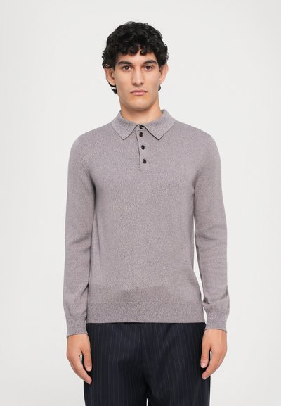 Junger Mann mit lockigem Haar, der einen helllilanen, gestrickten Polopullover mit Knöpfen und dunkel gestreifte Hosen trägt und vor einem schlichten Hintergrund steht.