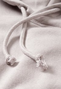 Gros plan sur un tissu de sweat à capuche beige clair avec deux cordons tressés terminant par des nœuds effilochés.