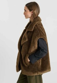 Bruine fleece vest met een kraag, twee zijzakken en een losse pasvorm. Heeft een zachte textuur en wordt gedragen over een zwart longsleeve shirt.