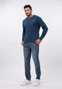 Mėlynas megztinis su pynimo raštais ir ketvirčio užtrauktuku apykaklėje, derinamas su šviesiai mėlynomis suplyšusiomis džinsinėmis kelnėmis ir baltais sportbačiais.