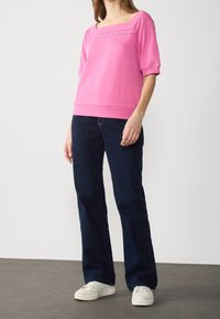 Žena oblečená v růžovém topu s krátkým rukávem a čtvercovým výstřihem, tmavě modrých džínách a bílých teniskách, stojí na šedé podlaze před bílou zdí.