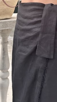 Falda negra de tela envolvente con cinturilla anudada y detalles deshilachados, usada junto a un balaustrada de piedra blanca y una bolsa de paja tejida.