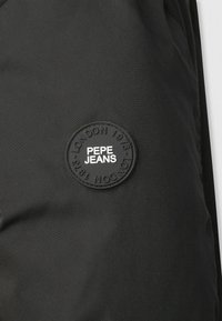 Černá textilie s výrazným kulatým černým gumovým nášivkem, který obsahuje vylisovaný bílý nápis "PEPE JEANS" a "LONDON 1973".