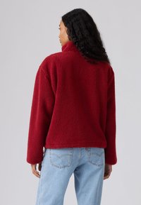 Camisola vermelha de lã com gola alta e superfície texturada e felpuda. Corte quadrado, ombros caídos e mangas compridas combinadas com jeans azul claro.