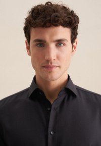 Camicia nera con bottoni, con un motivo testurizzato, colletto aperto e maniche a sbuffo. Il modello ha i capelli corti e ricci e occhi azzurri.