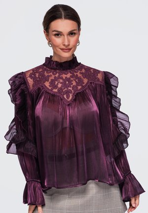 Femme portant un chemisier violet transparent avec un empiècement en dentelle florale et des manches à volants, assorti à un pantalon clair à carreaux, souriante sur un fond uni.