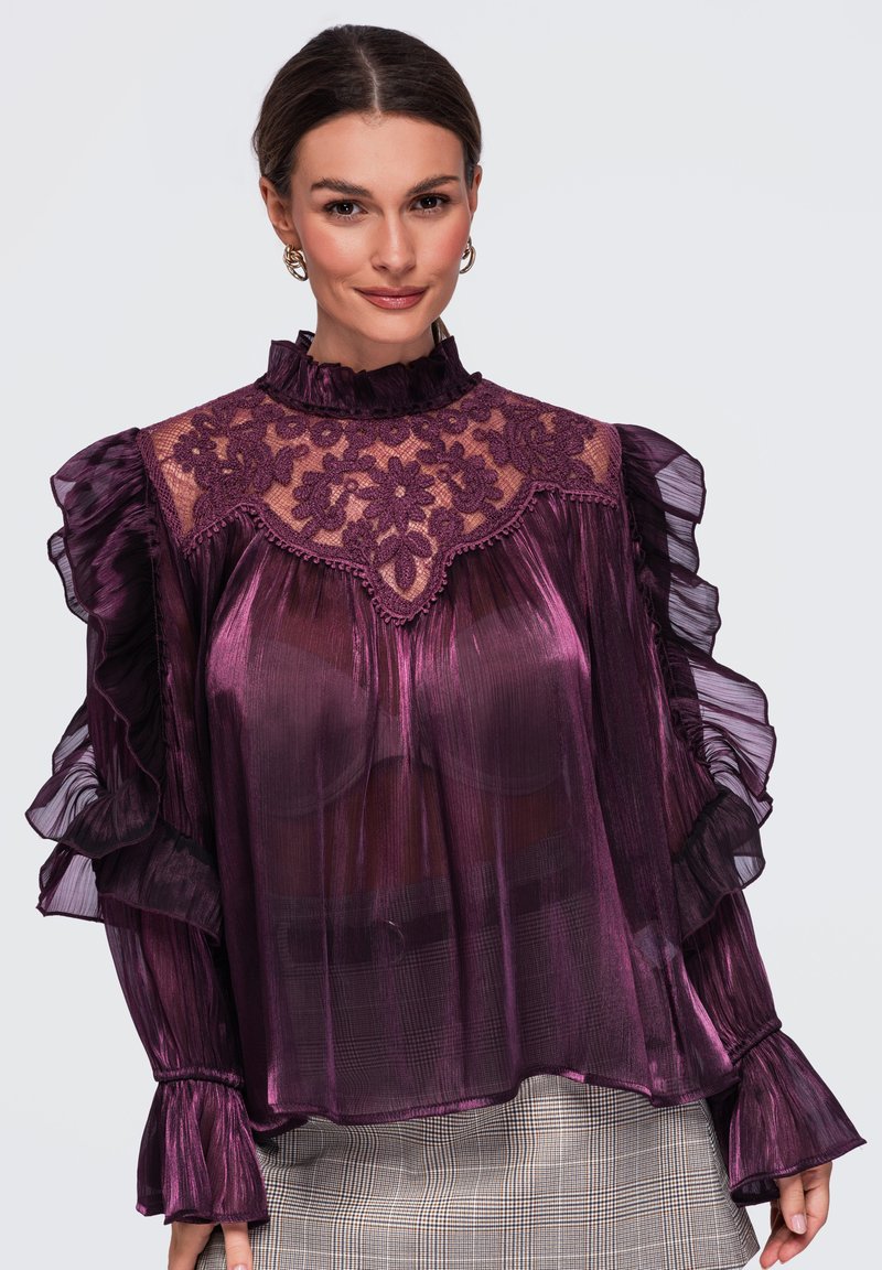 Femme portant un chemisier violet transparent avec un empiècement en dentelle florale et des manches à volants, assorti à un pantalon clair à carreaux, souriante sur un fond uni.