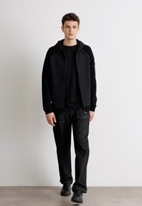 Črna kapucar na zadrgo s sprednjimi žepi, nošena preko črne majice, v kombinaciji z temnimi teksturiranimi hlačami in črnimi športnimi copati.