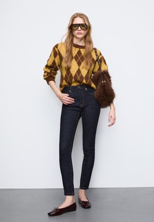 Brun och gul argyle-tröja, mörkblå skinny jeans, en axelväska i päls, gröna solglasögon och mörka platta skor. Släta texturer och figurnära design.