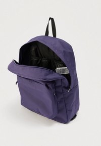 Sac à dos violet avec sangle noire, poche zippée à l'avant et compartiment principal partiellement ouvert montrant la doublure intérieure noire et une étiquette.