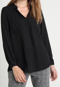 Blusa nera a maniche lunghe con scollatura a V, due tasche sul petto e polsini abbottonati. Il tessuto ha una superficie liscia. Abbinata a pantaloni a scacchi.