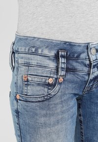 Džínové kalhoty ve světle modré barvě s vybledlým vzhledem, s měděnými doplňky, pěti kapsami a prošíváním podél pasu a kapes.