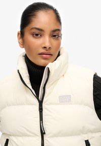 Gilet imbottito color crema con collo alto e cerniere. Presenta una toppa con il logo Superdry sul petto sinistro e un design trapuntato.