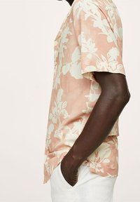 Personne à la peau foncée portant une chemise à manches courtes rose clair avec un motif floral blanc et un pantalon blanc, les mains dans les poches.