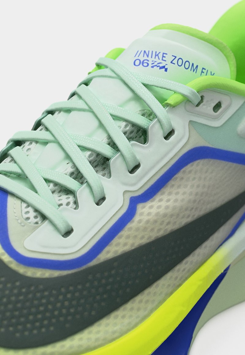 Gros plan sur une chaussure de course Nike Zoom Fly 4 vert clair avec des accents bleus, gris foncé et jaune néon, mettant en avant les lacets et la languette.