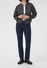 Femme portant une veste zippée gris foncé sur une chemise blanche, un jean bleu foncé et des mocassins noirs à semelles épaisses, debout devant un fond blanc.