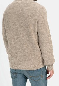 Beiger gestrickter Pullover mit strukturiertem Finish, Rundhalsausschnitt und langen Ärmeln, kombiniert mit blauen Jeans aus Denim, die über Rückentaschen mit Stickdetails verfügen.