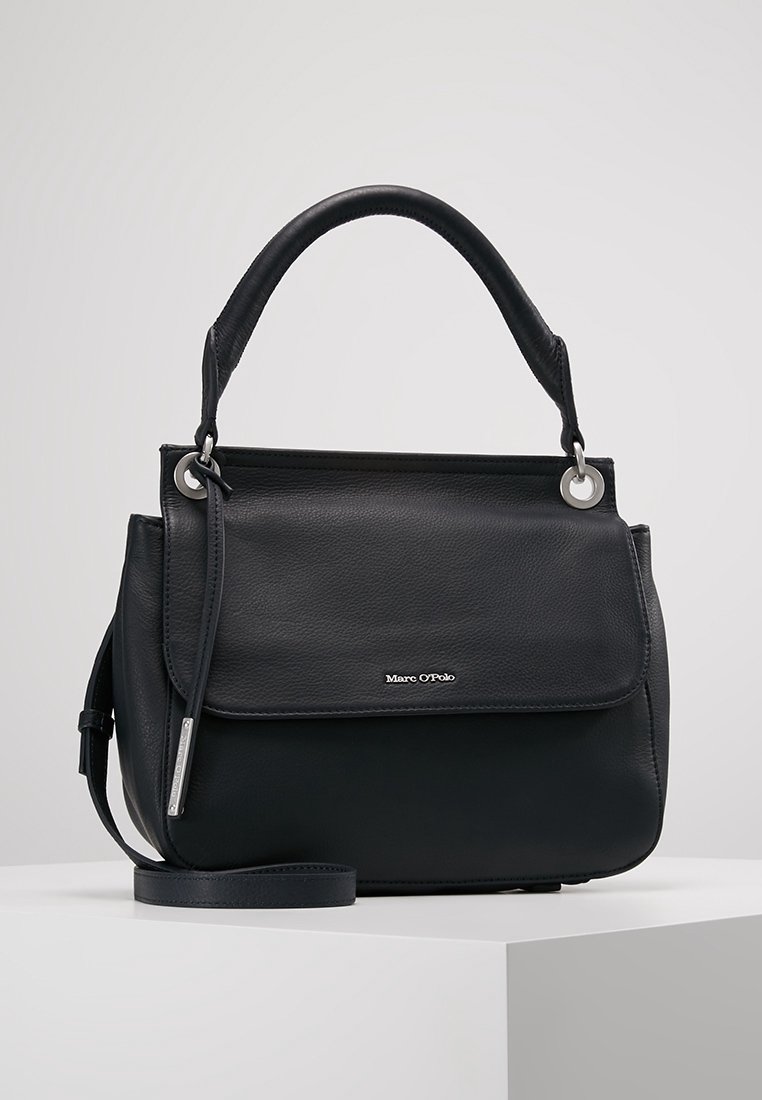 Sac à main en cuir noir Marc O’Polo avec poignée supérieure, rabat avant et bandoulière amovible, posé sur une surface blanche.