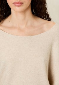 Žena s kučeravými tmavými vlasmi nosí béžový pletený sveter s odhalenými ramenami, ktorý ukazuje výstrih a hornú časť hrude na neutrálnom pozadí.