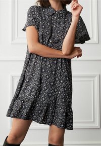 Kvinde i en kortærmet, knaplukket kjole med et sort-hvidt geometrisk mønster, der står med krydsede arme mod en hvid panelvæg.