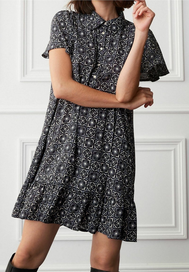 Kvinde i en kortærmet, knaplukket kjole med et sort-hvidt geometrisk mønster, der står med krydsede arme mod en hvid panelvæg.