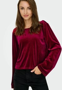 Karmosinröd sammetströja med lång ärm och fyrkantig halsringning, avslappnad passform och samlade axeldetaljer, kombinerad med svarta jeans.