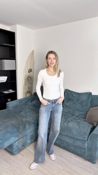 Camiseta blanca de manga larga combinada con jeans acampanados de color azul desgastado y zapatos blancos. Sofá de terciopelo verde azulado y una tabla de surf con un diseño desgastado.