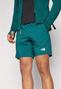 Shorts légers teal en tissu lisse. Présente un logo blanc et une poche latérale avec fermeture éclair. Conçu pour les activités sportives.