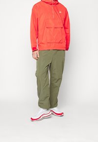 Röd vindjacka med ficka fram, gröna löst sittande byxor och vita sneakers med röda accenter, med en elegant design och andningsbara material.