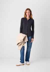 Marineblå skjorte med hvite prikker, lange ermer, kombinert med lyseblå jeans og holder en kremfarget jakke.