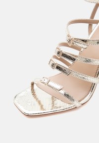 Μια μεταλλική χρυσή σανδάλι με πέντε ιμάντες με ρυθμιζόμενες αγκράφες και τετράγωνη μυτερή μύτη, με υφή λαμπερή και λαμπερό φινίρισμα.