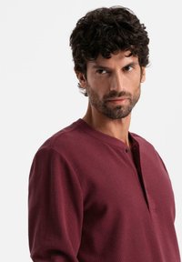 Uomo con capelli ricci e scuri e barba, che indossa una maglietta henley a maniche lunghe di colore bordeaux, mentre guarda leggermente di lato su uno sfondo bianco semplice.